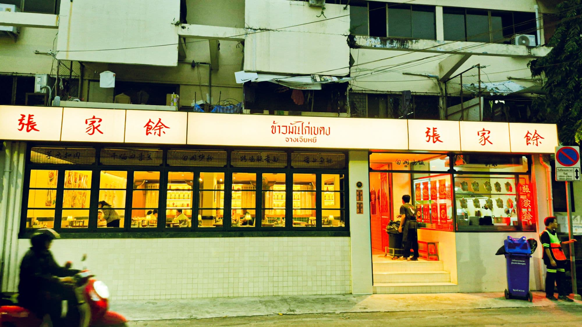 A restaurant front at dusk, its signs lit in Chinese and Thai, a scooter blurring past