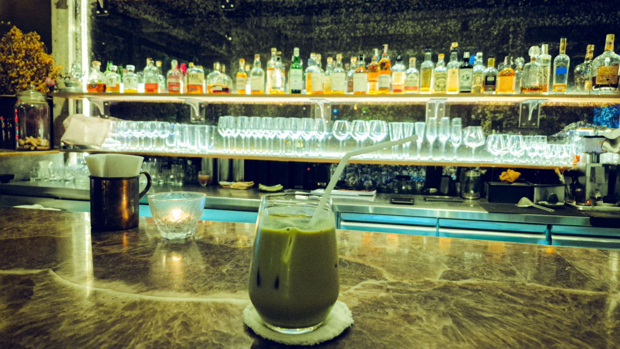 An iced green drink with a straw on a marble bar, rows of bottles and glasses behind