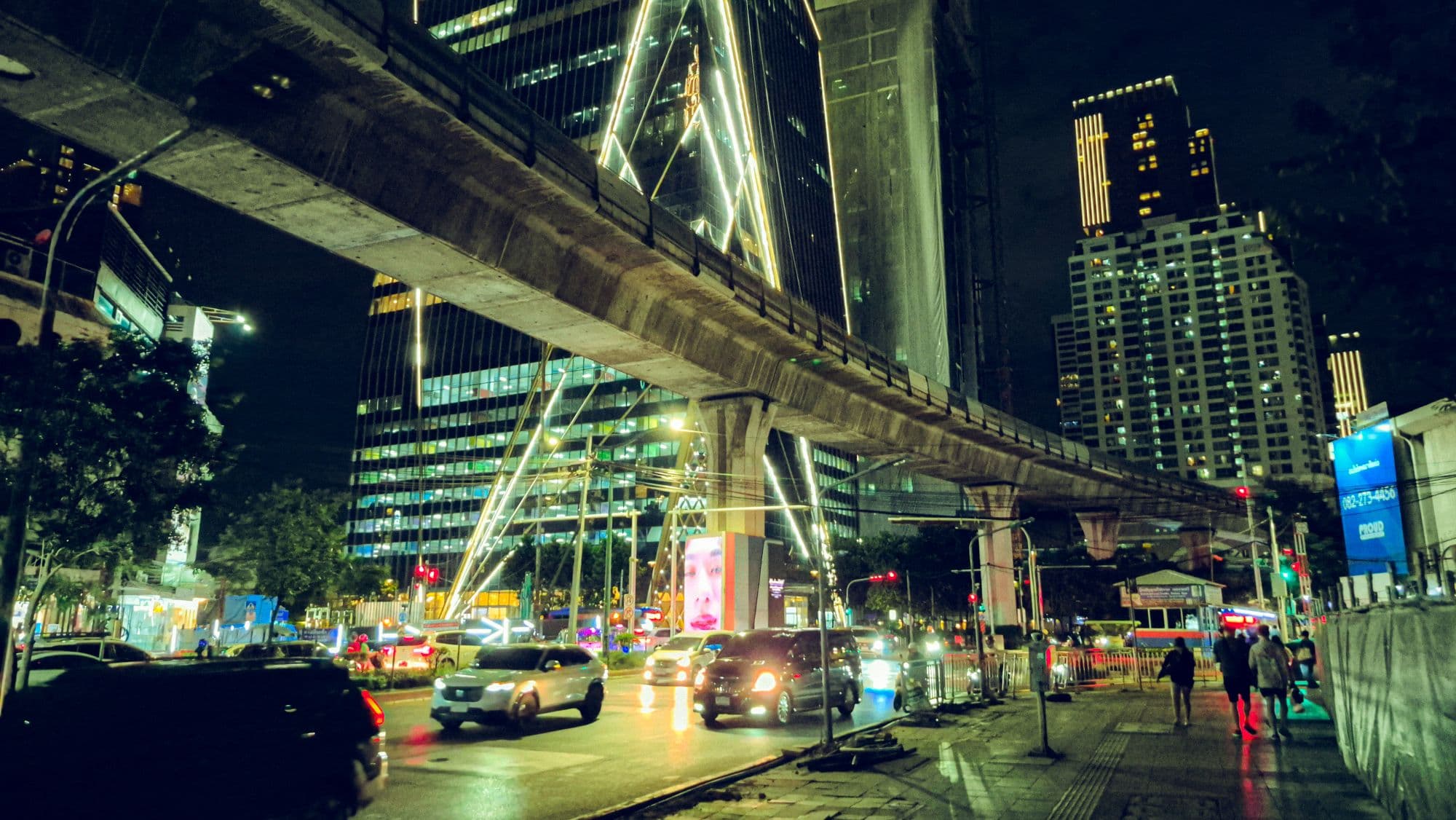 A raised train line crossing a busy street at night, lit towers behind it