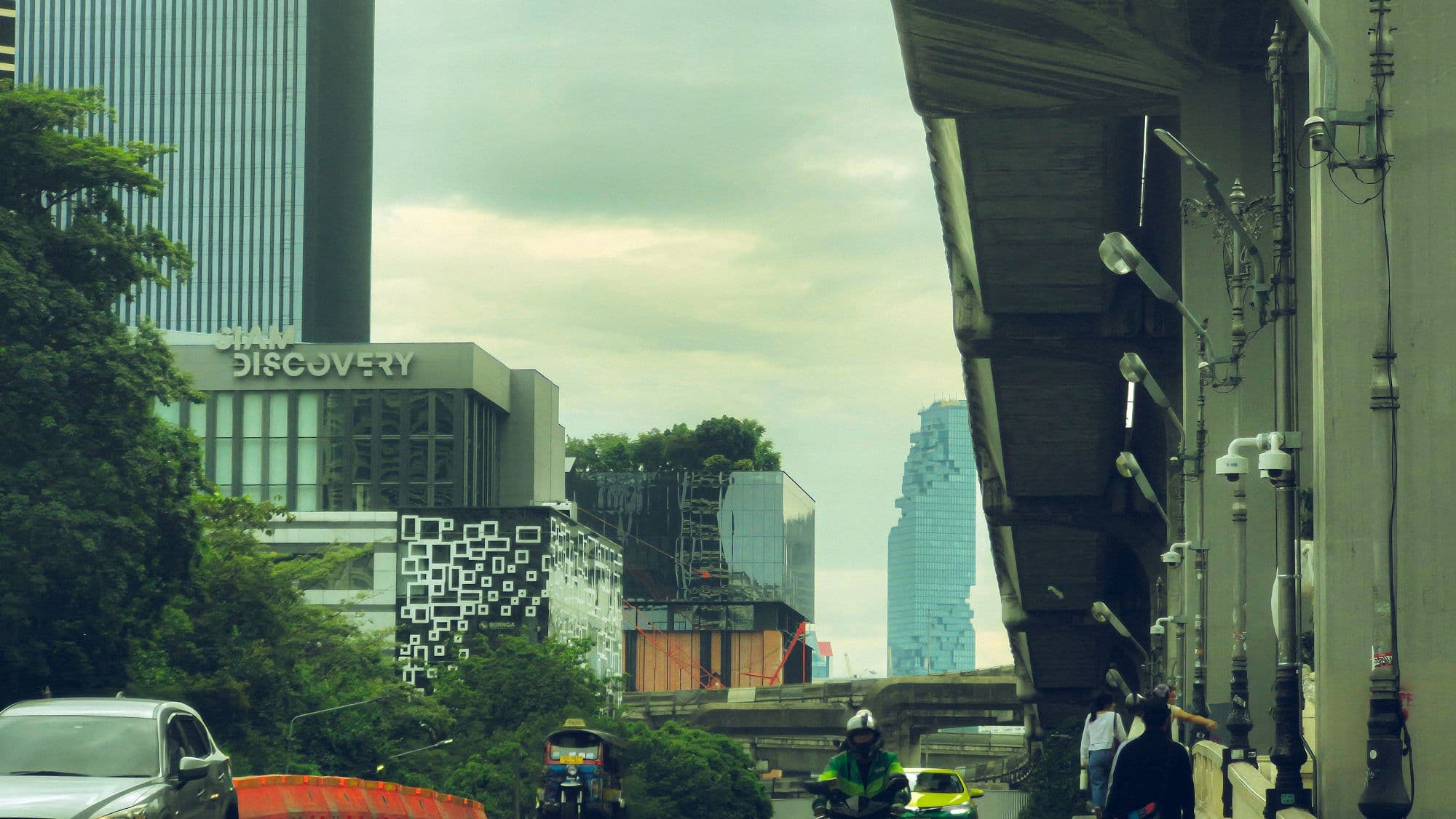 A raised train line along a street, with a glass tower and low buildings beyond