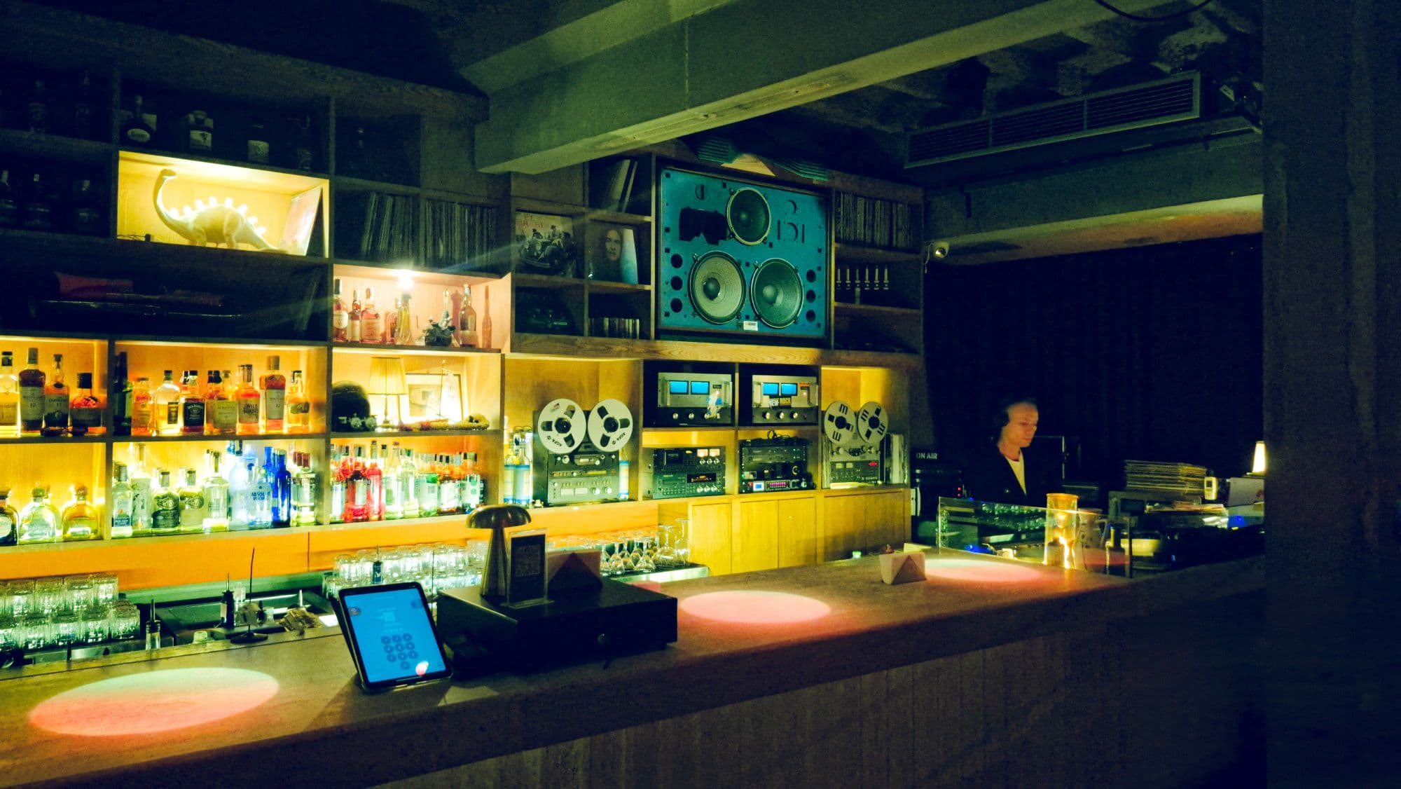 A bar lit amber, with a speaker, tape machines and a person in headphones behind the counter