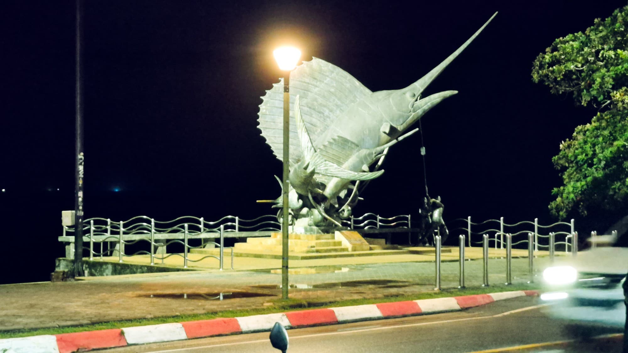 A large sailfish sculpture on a seafront at night, under a lamp