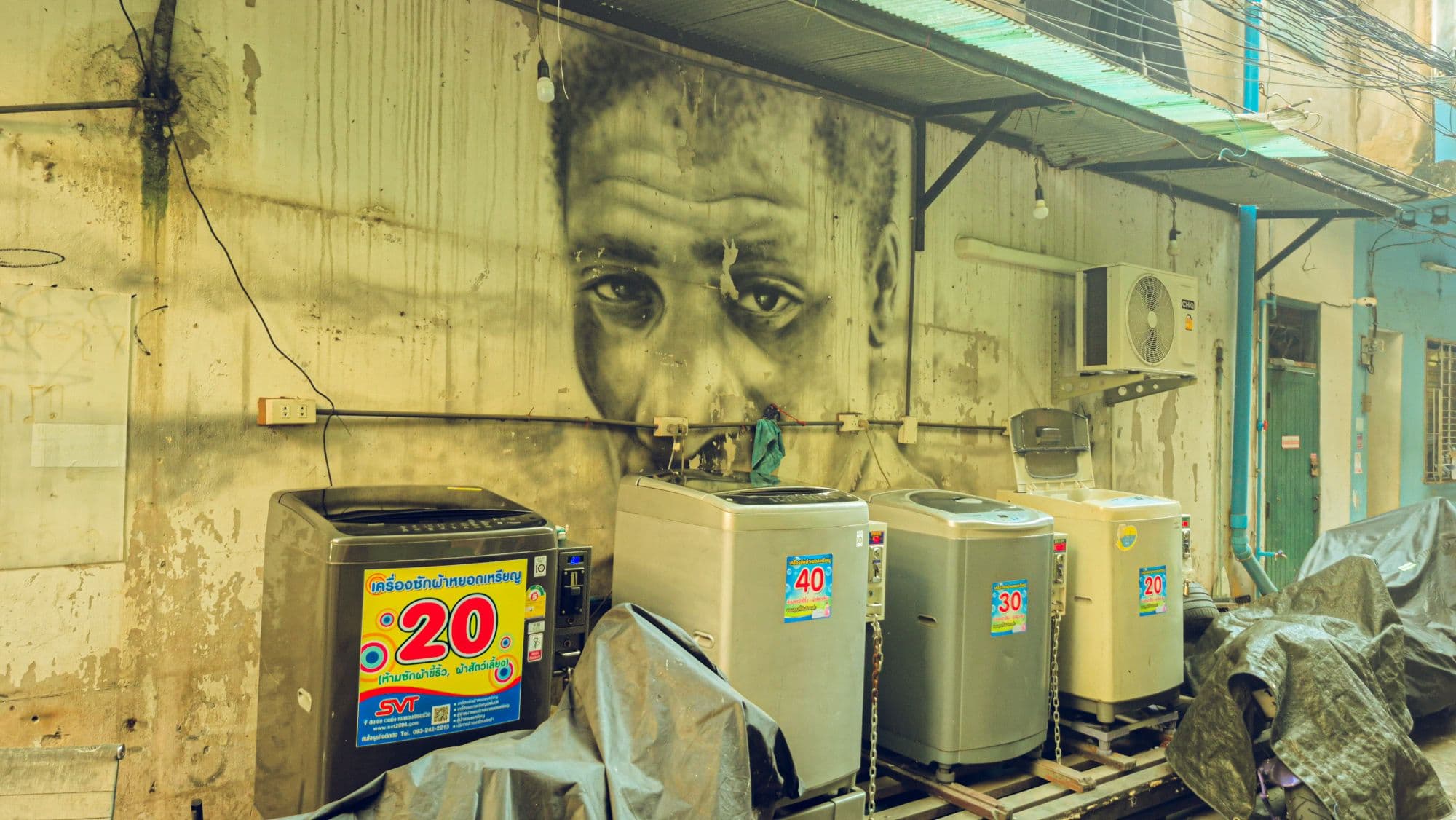 A large painted face on a weathered wall above four washing machines